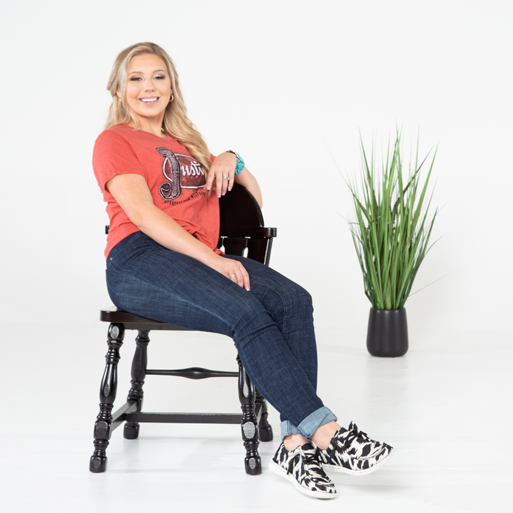A blonde woman sitting in a chair leaned back next to a green plant, showcasing her cowprint casual shoes.